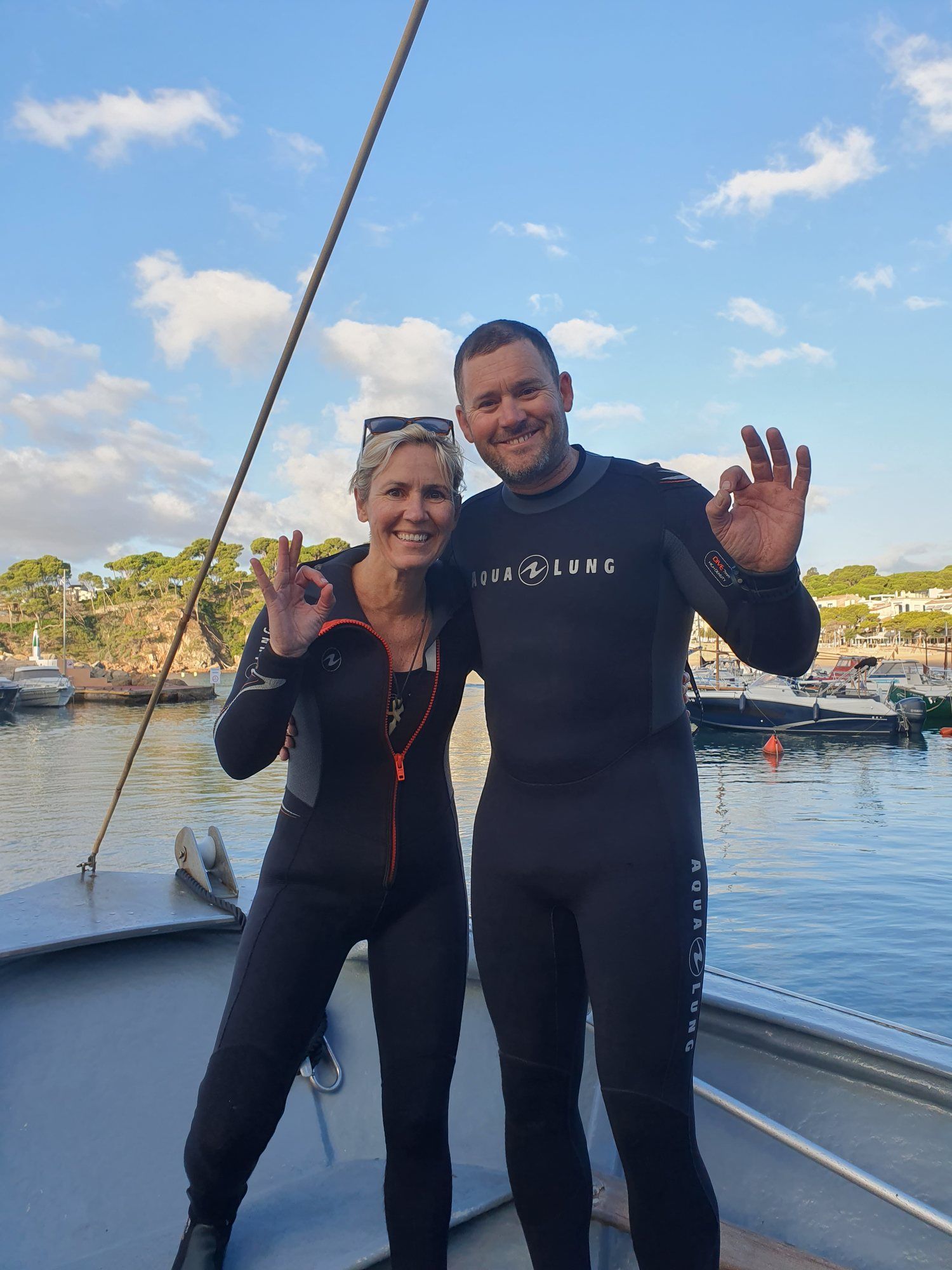 Mim and Mark in wetsuits on a boat, both giving the OK sign